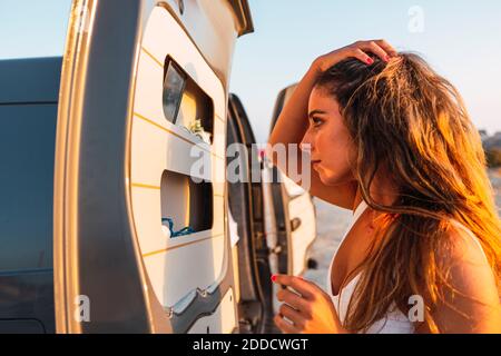 Junge Frau mit der Hand im Haar und Blick auf den Spiegel in Wohnmobil Tür beim Stehen am Strand Stockfoto