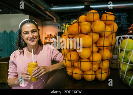 Lächelnd reife Frau hält frischen Saft in Einmachglas, während Stehen bei Orangen im Metallkorb im Café Stockfoto