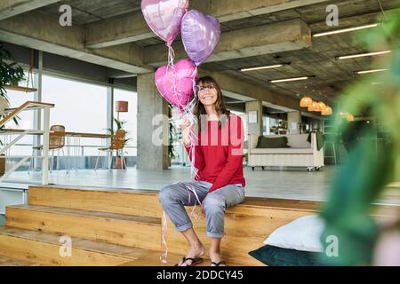 Geschäftsfrau hält Ballons, während sie auf Treppen im Loft-Büro sitzt Stockfoto