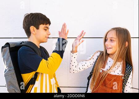 Bruder und Schwester tun hohe fünf, während sie gegen Weiß stehen Wand an sonnigen Tag Stockfoto