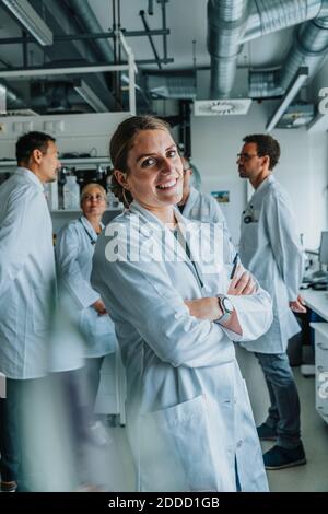 Junger Wissenschaftler lächelt, während er mit gekreuzten Armen und Mitarbeiter steht Im Hintergrund im Labor Stockfoto