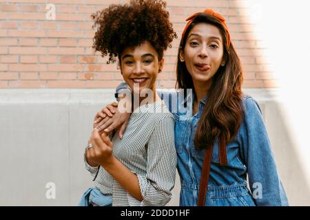 Junge Frau, die die Zunge herausstreckt, während sie mit einem Freund dagegen steht Wand Stockfoto