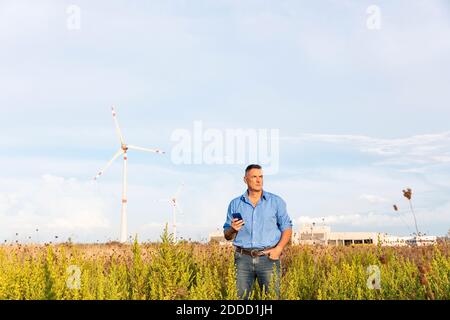 Reifer Geschäftsmann hält Smartphone, während er gegen Windturbinen steht Auf dem Feld Stockfoto