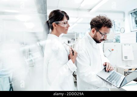 Eine Wissenschaftlerin steht bei einem Kollegen, der im Labor einen Laptop benutzt Stockfoto