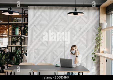 Junge Frau trägt schützende Gesichtsmaske sprechen auf Smartphone Während der Verwendung von Laptop im Café Stockfoto