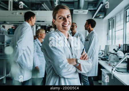 Lächelnde Frau mit gekreuzten Armen stehend mit Mitarbeiter im Hintergrund Im Labor Stockfoto