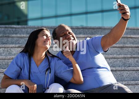 Lächelnde Ärzte, die Selfie machen, während sie auf der Treppe gegen das Krankenhaus sitzen Stockfoto