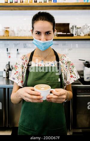 Junge Frau trägt Gesichtsmaske und Schürze mit Kaffeetasse Im Café stehen Stockfoto