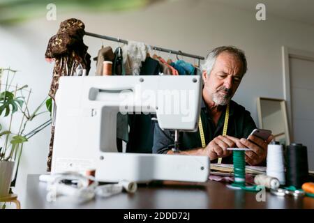 Reifer männlicher Schneider mit Smartphone, während er am Tisch sitzt Im Arbeitsstudio Stockfoto