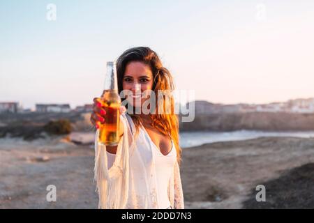 Lächelnde Frau, die während des Strands eine kleine Weißweinflasche zeigt Sonnenuntergang Stockfoto