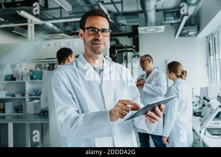 Lächelnder Wissenschaftler mit digitalem Tablet, während er mit einem Kollegen steht Im Hintergrund im Labor Stockfoto