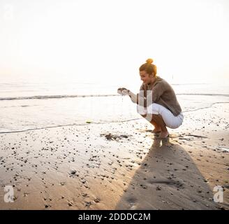 Schöne junge Frau hocken, während sie Sand am Ufer hält Strand bei Sonnenuntergang Stockfoto