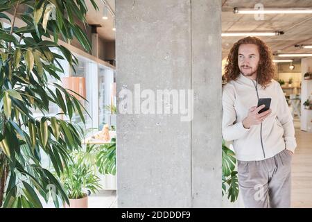 Mann mit der Hand in der Tasche hält Smartphone, während lehnte Auf grauer Säule zu Hause Stockfoto