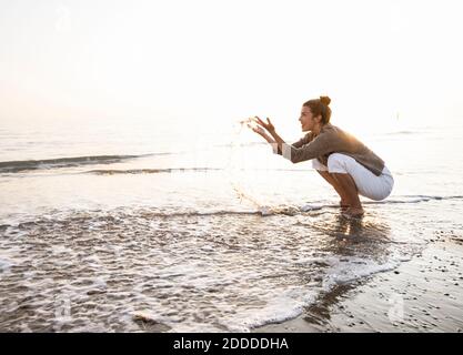 Schöne junge Frau, die während des Strands Wasser am Ufer spritzt Sonnenuntergang Stockfoto