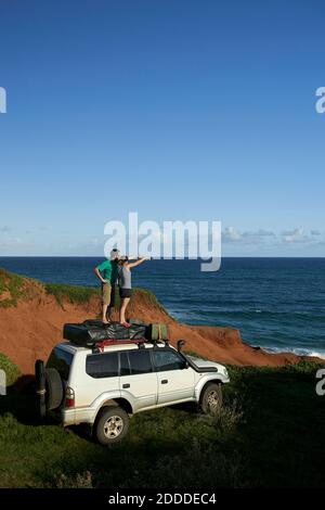 Paar zeigt, während man auf dem 4x4 Dach steht, während man anschaut Meer gegen blauen Himmel Stockfoto