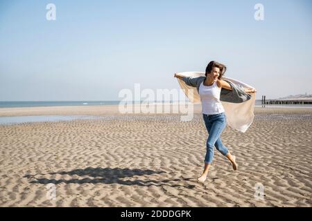 Sorglose junge Frau läuft, während sie am Strand eine Decke hält Der Himmel ist klar Stockfoto