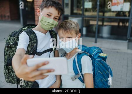 Brüder, die Selfie auf dem Smartphone machen, während sie ein schützendes Gesicht tragen Maske steht vor dem Schulgebäude Stockfoto
