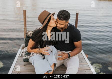 Frau mit Weinglas küssen Mann, während auf Pier sitzen Stockfoto