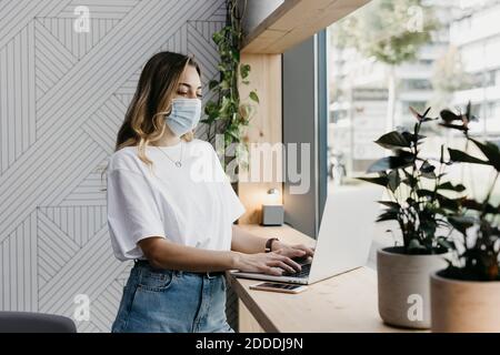 Frau trägt eine schützende Gesichtsmaske mit Laptop, während sie im Stehen ist Cafe während Covid-19 Stockfoto