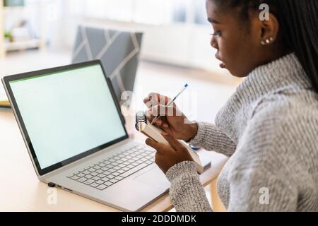 Junge Frau schreiben in Notepad während studieren durch Laptop an Zu Hause Stockfoto