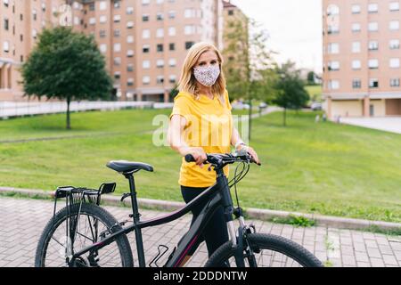 Reife Frau trägt schützende Gesichtsmaske stehen mit Fahrrad auf Fußweg in der Stadt während der COVID-19 Stockfoto