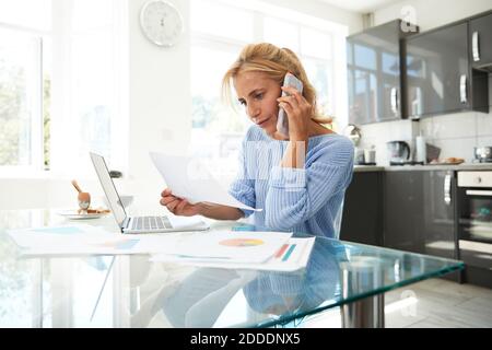 Reife Frau am Telefon sprechen, während in der Küche zu Hause Stockfoto