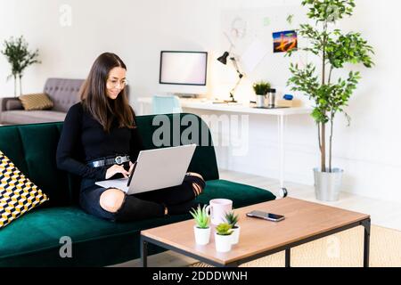 Lächelnd schöne junge Frau mit Laptop zum Bloggen während des Sitzens Auf dem Sofa zu Hause