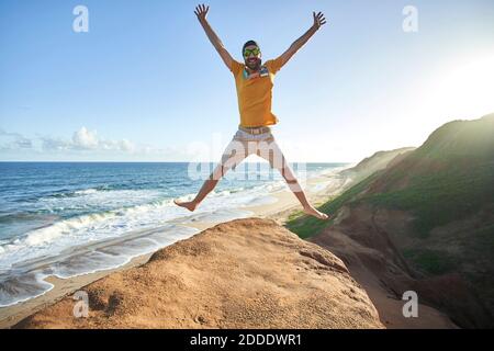 Fröhlicher Mann, der am Strand gegen Blau auf Felsformation springt Himmel Stockfoto