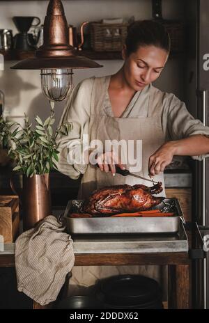 Junge Frau in Leinenschürze Carving ganze geröstete Ente für Thanksgiving-Urlaub oder Heiligabend Party Feier in modernen Küche Interieur. Traditionelles Komfortessen im Herbst oder Winter Stockfoto