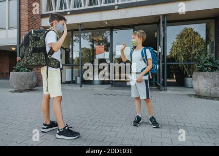 Brüder winken, während sie eine Gesichtsmaske tragen, die davor steht Des Schulgebäudes Stockfoto