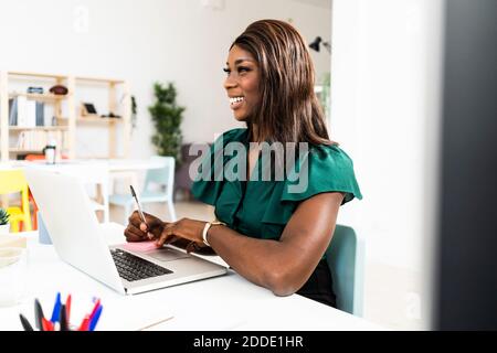 Lächelnde Geschäftsfrau schreiben auf Klebenote während der Arbeit auf Laptop Im Büro Stockfoto