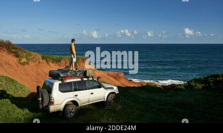 Männlicher Tourist, der auf dem 4x4-Dach steht und das Meer betrachtet Am blauen Himmel Stockfoto