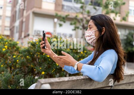 Frau gestikuliert, während mit Smartphone auf Bank in der Stadt Während COVID-19 Stockfoto