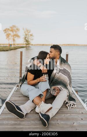 Mann küsst Frau, während auf Pier gegen See sitzen Stockfoto