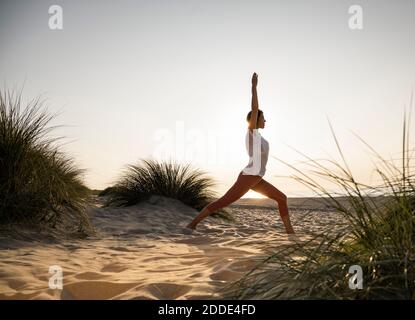 Junge Frau praktiziert Kriegerposition Yoga inmitten von Pflanzen am Strand Gegen klaren Himmel bei Sonnenuntergang Stockfoto