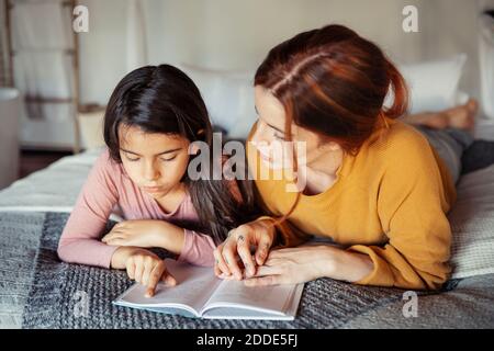 Tochter beim Lesen Buch, während sie auf dem Bett von der Mutter liegt Zu Hause Stockfoto