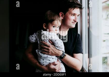 Lächelnder Vater, der Sohn trägt, während er durch das Fenster schaut Zu Hause Stockfoto