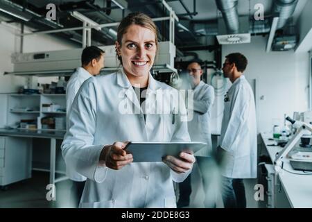 Lächelnde Frau mit digitalem Tablet, während sie mit einem Kollegen im Stehen steht Hintergrund im Labor Stockfoto