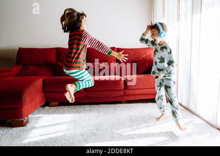 Männliche und weibliche Geschwister tragen Kopfhörer, während sie gegen das Sofa tanzen Im Wohnzimmer Stockfoto