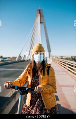 Frau in schützende Gesichtsmaske mit Elektroroller auf Brücke In der Stadt während der COVID-19 Stockfoto