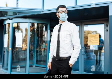 Mann Geschäftsmann in Business-Kleidung und eine medizinische Schutzmaske auf seinem Gesicht, vor dem Hintergrund eines Glas-Bürogebäude Stockfoto