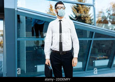 Mann Geschäftsmann in Business-Kleidung und eine medizinische Schutzmaske auf seinem Gesicht, vor dem Hintergrund eines Glas-Bürogebäude Stockfoto