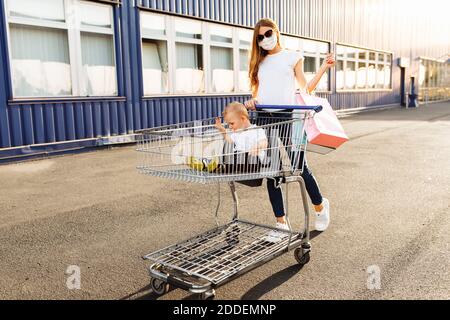 Glückliche junge Familie, Mama in medizinischer Maske mit Taschen und lächelndes Kind sitzt in einem Trolley, Familie geht einkaufen Stockfoto