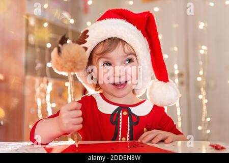 Kleines glückliches Mädchen in einem roten Kleid und Hut schreibt einen Brief an den Weihnachtsmann. Das Kind wünscht sich Weihnachten. Das Baby lächelt, schreibt mit einem Stift auf einem Stockfoto