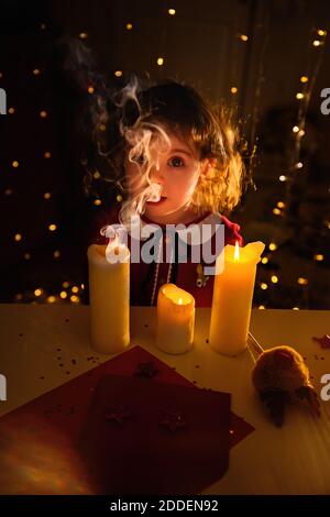 Neujahrs-Porträt eines kleinen schönen Mädchen in einem roten Kleid. Das Kind wünscht sich Weihnachten. Das Baby bläst die Kerzen aus. Verschwommenes Backgroun Stockfoto