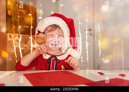 Kleines glückliches Mädchen in einem roten Kleid und Hut schreibt einen Brief an den Weihnachtsmann. Das Kind wünscht sich Weihnachten. Das Baby schloss die Augen, schreibt mit einem Stockfoto