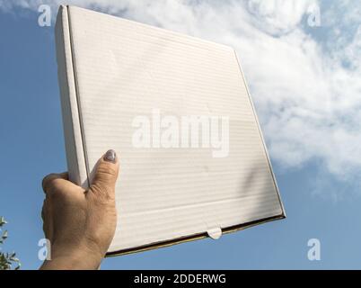 Eine Frauenhand hält eine weiße Pappschachtel mit einer Kopie des Raumes und des Platzes für Text auf einem blauen Himmel Hintergrund, Schablone, mockup Stockfoto