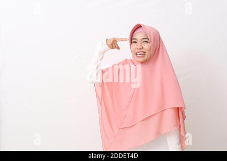 Ein Porträt asiatische muslimische Frau trägt einen rosa Schleier oder Hijab und weißes Kleid gestikulieren mit einem Finger gegen Tempel sagen, Sie sind verrückt. Zeigt auf h Stockfoto