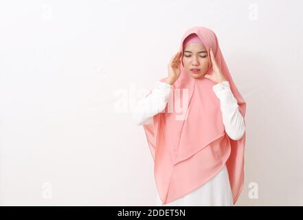 Ein Porträt einer asiatischen muslimischen Frau mit einem rosa Schleier oder Hijab und weißem Kleid bekam Kopfschmerzen. Sie berührte ihre Kopfbügeln mit beiden Händen und Leid Stockfoto