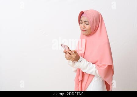 Ein Porträt einer traurigen asiatischen muslimischen Frau, die einen Schleier oder Hijab mit einem Handy trägt und weint. Schlechte Nachrichten erhalten. Isoliert auf weißem Hintergrund mit Kopie Stockfoto